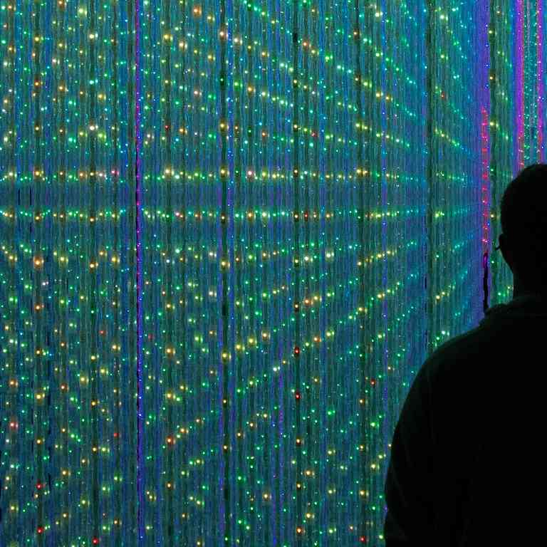 Silhouetted person standing inside an immersive light installation, surrounded by dense rows of blue, turquoise, green and purple lights reflected across the space.
